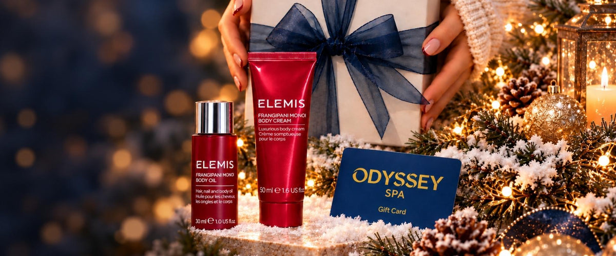 A festive holiday gift scene featuring two red Elemis skincare bottles and a blue Odyssey Spa gift card, arranged on a snowy decorated surface with lights, pinecones, ribbons, and a gift box in the background.