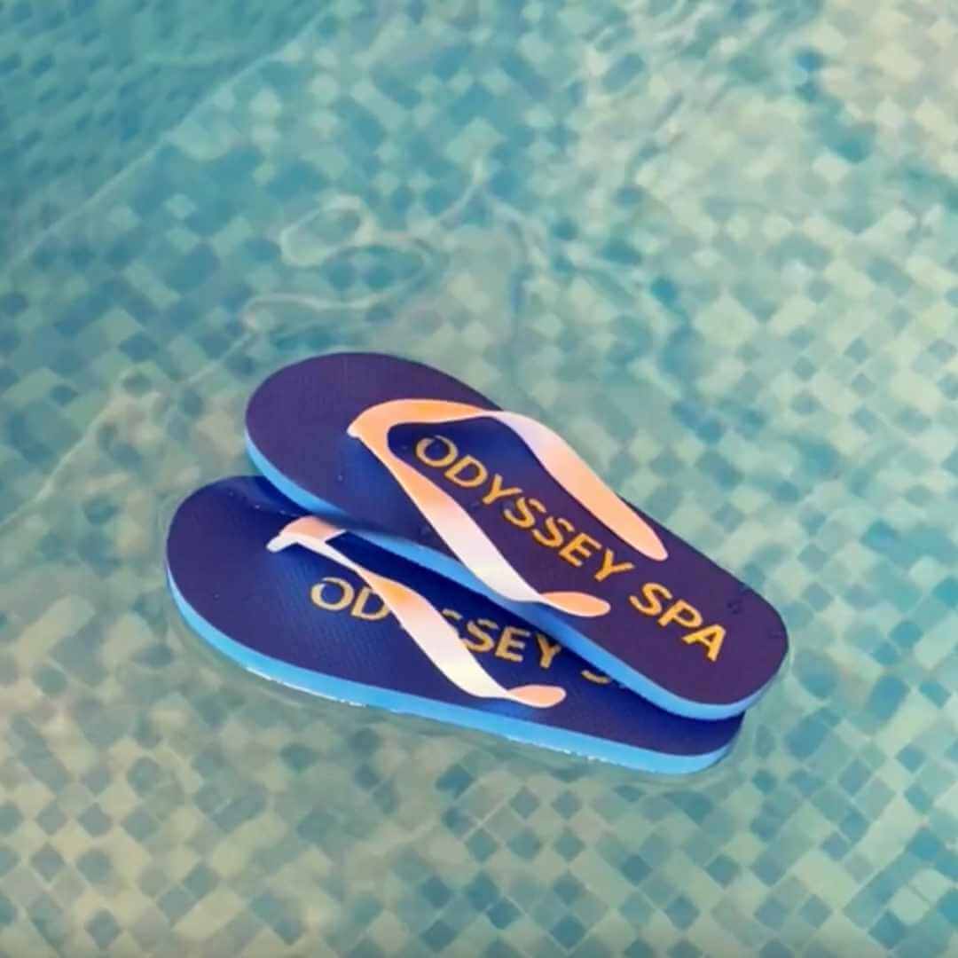 A pair of blue Odyssey branded flip flops floating on a pool, with the Odyssey Spa logo visible on the top.