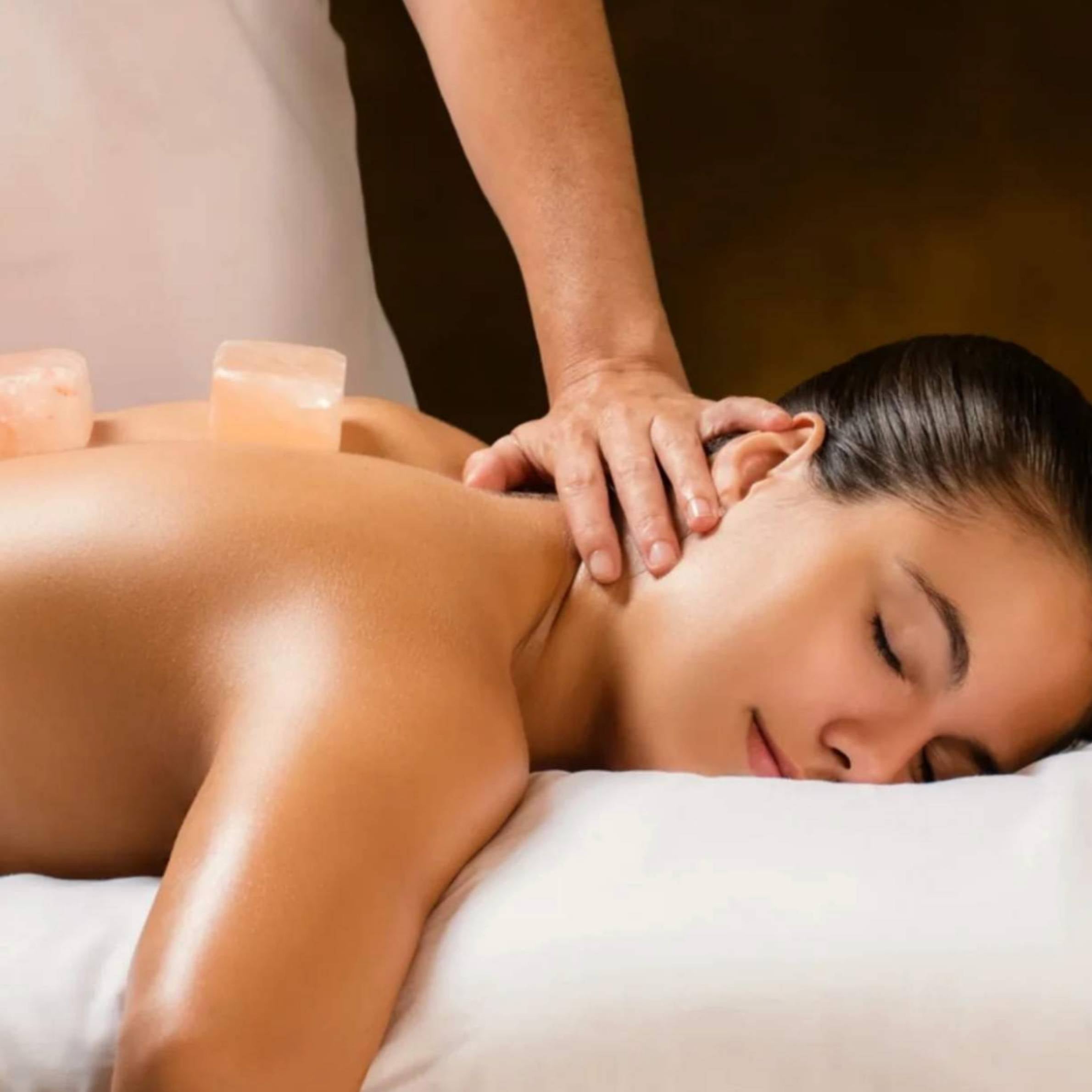 A woman lies face down on a massage table while a therapist massages her neck and upper back. Salt candles are visible on the table.