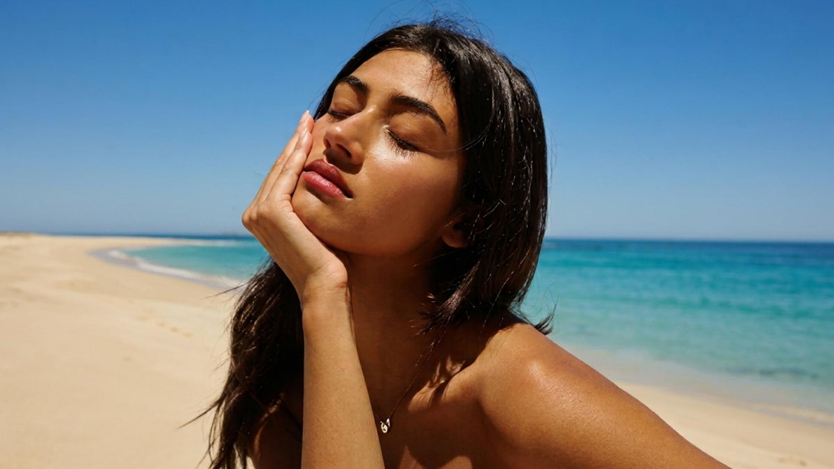 A woman holding her glowing face against the backdrop of a sandy beach and blue sea