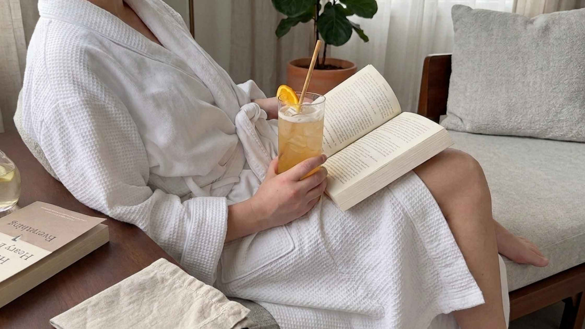 A woman sitting in a white spa robe whilst reading a book and holding an orange drink