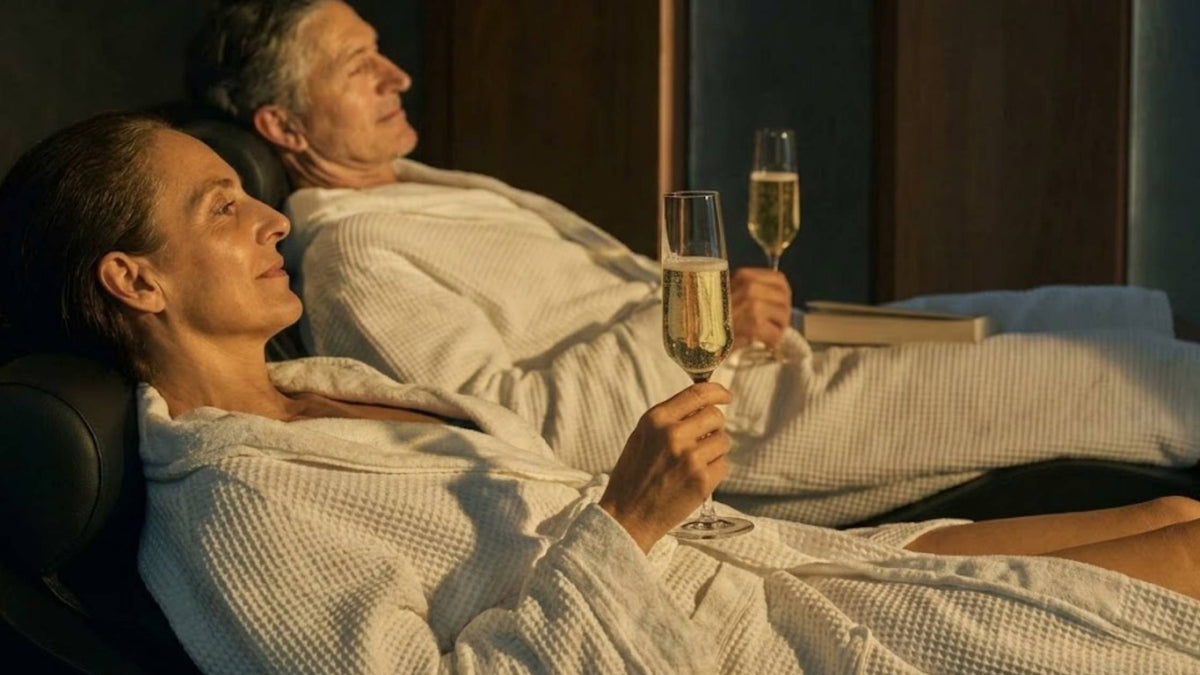 Two women in white robes laying down and enjoying a glass of prosecco
