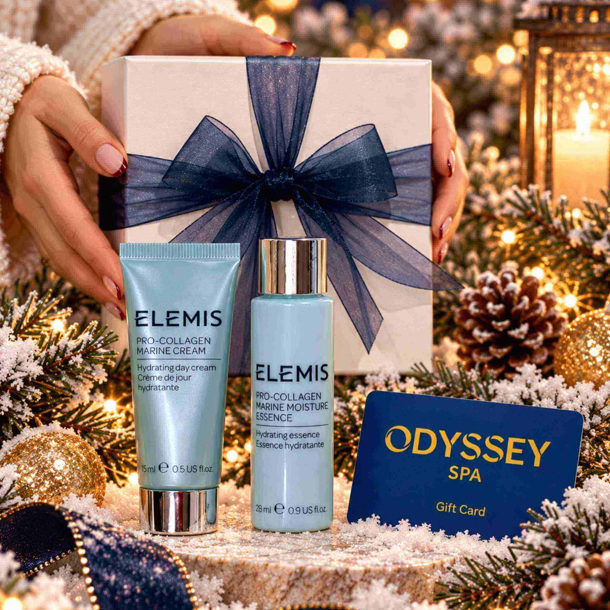 A gift set featuring two ELEMIS skincare products (Pro-Collagen Marine Cream tube and Bio-Collagen Marine Moisture Essence bottle) in light blue packaging with silver caps, presented in front of a white gift box with a navy blue bow. The set is held by hands wearing a white knit sweater, surrounded by festive Christmas decorations including pine branches, gold ornaments, a candle, and an Odyssey Spa gift card.