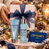 A gift set featuring two ELEMIS skincare products (Pro-Collagen Marine Cream tube and Bio-Collagen Marine Moisture Essence bottle) in light blue packaging with silver caps, presented in front of a white gift box with a navy blue bow. The set is held by hands wearing a white knit sweater, surrounded by festive Christmas decorations including pine branches, gold ornaments, a candle, and an Odyssey Spa gift card.