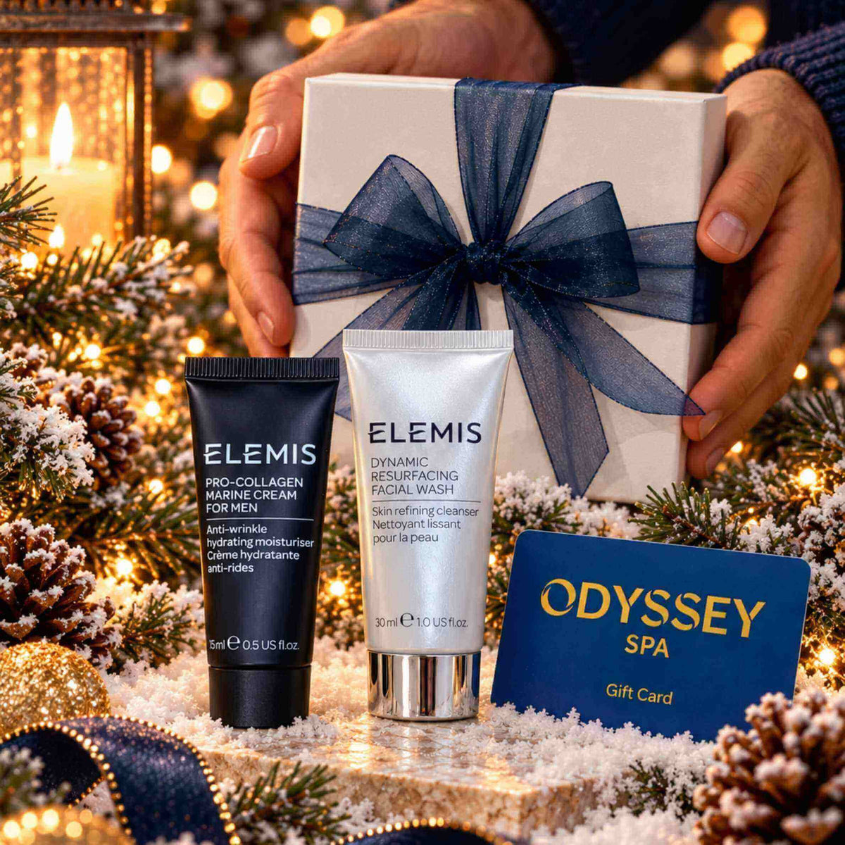 A festive gift set featuring two ELEMIS skincare products (a black tube and a white bottle) arranged on a snowy, decorated Christmas tree branch next to an Odyssey Spa gift card, with a white gift box tied with a navy ribbon held by hands in the background.
