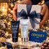 A festive gift set featuring two ELEMIS skincare products (a black tube and a white bottle) arranged on a snowy, decorated Christmas tree branch next to an Odyssey Spa gift card, with a white gift box tied with a navy ribbon held by hands in the background.