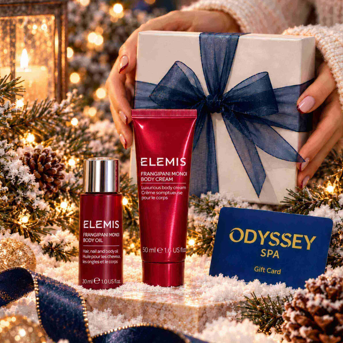 A festive gift set featuring ELEMIS Frangipani Mungi Body Cream in a red tube and ELEMIS Frangipani Mungi Body Oil in a red bottle with a silver cap, alongside an Odyssey Spa gift card, set against a snowy holiday backdrop with pine branches and a wrapped gift box held by hands.