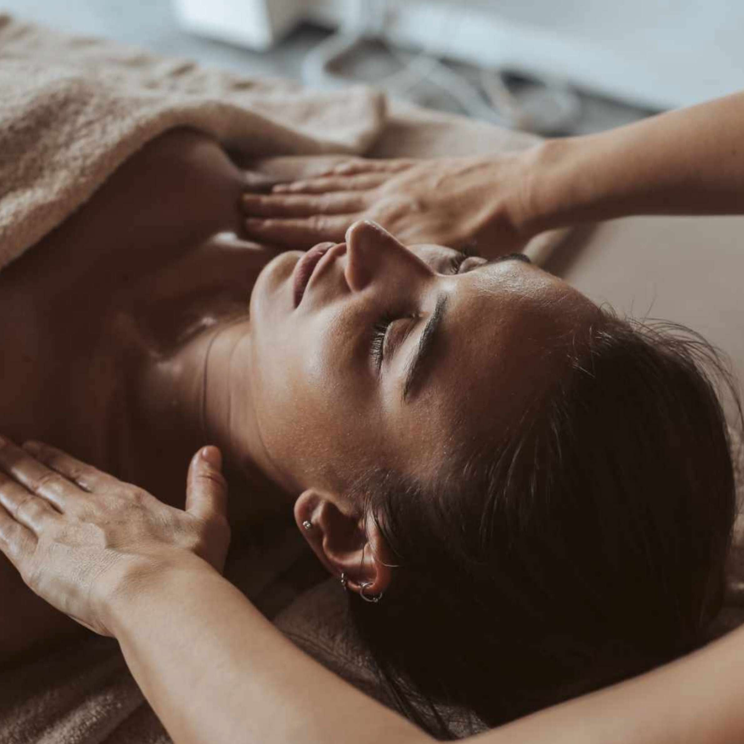 A close-up lifestyle shot of a woman receiving a massage, with her head resting on a towel-covered table while another person's hands massage her neck and chest.