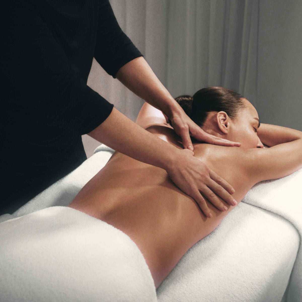 A person receiving a massage lies face down on a white treatment table while another person in a black top massages their upper back and shoulders in a spa-like setting.