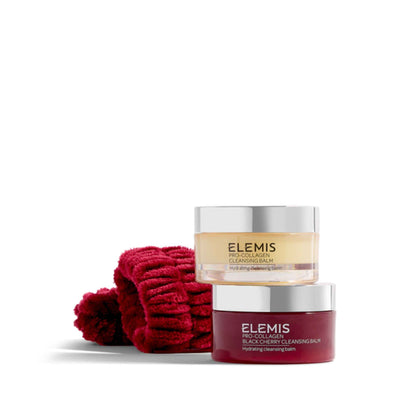 Two jars of Elemis Cleansing Balm with a folded red knit towel beside them on a clean white background. The front jar is dark red with a silver lid, and the back jar is pale yellow with a silver lid.